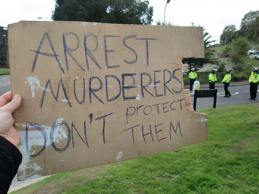 A placard held up against the police says "ARREST MURDERERS - DON'T PROTECT THEM"