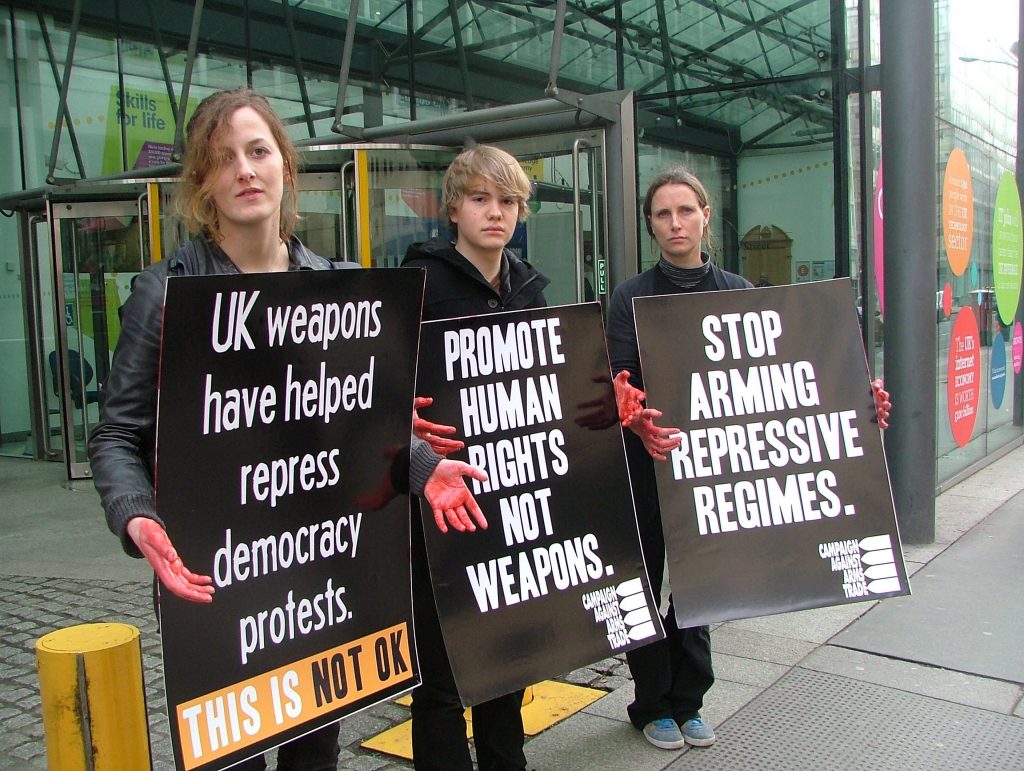 Three people outside a building. They have red hands and are carrying placards. One placard reads 'UK weapons have helped repress democracy protests'