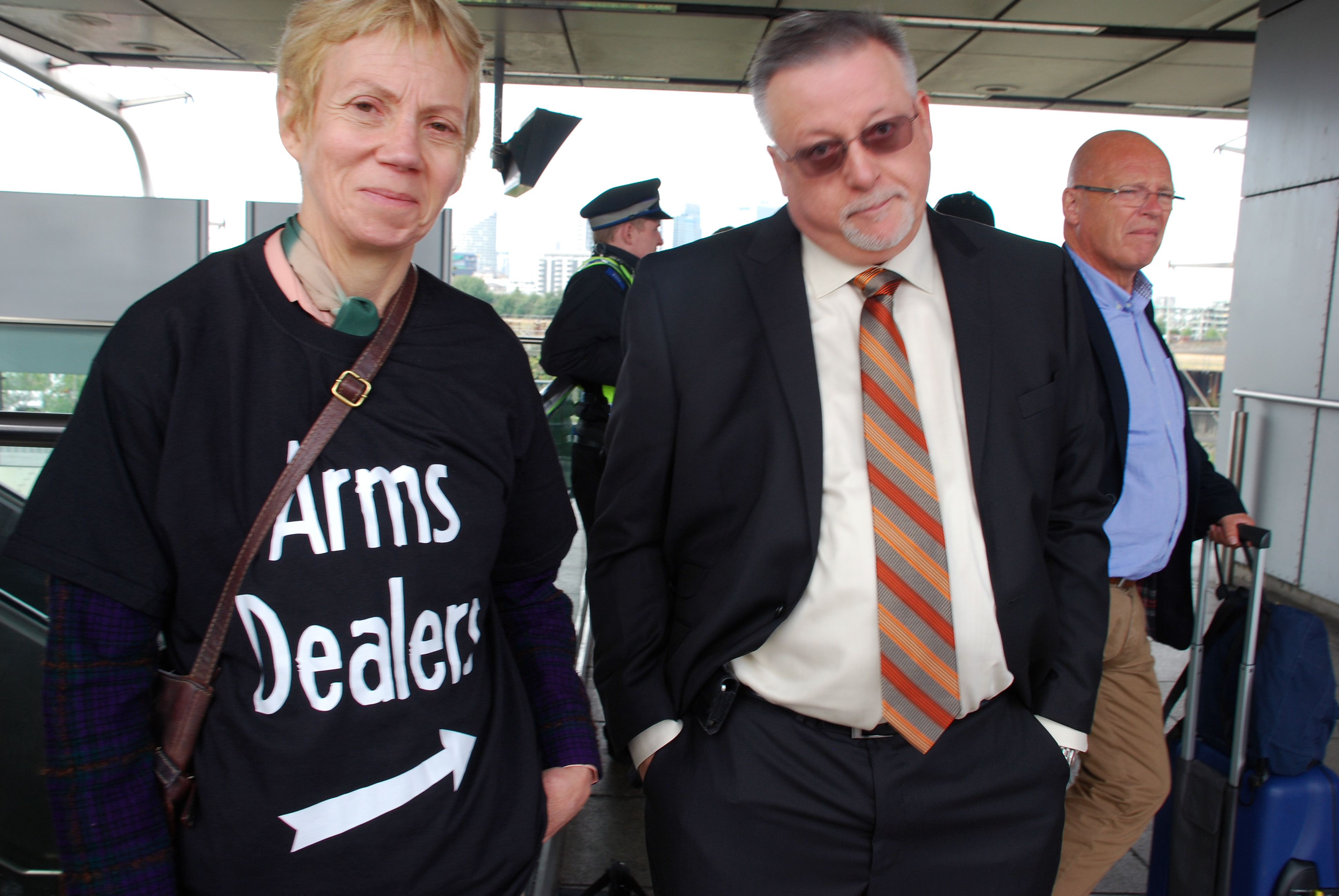 Activist with a T-shirt reading "Arms Dealer" and an arrow pointing sideways stands next to an arms fair delegate