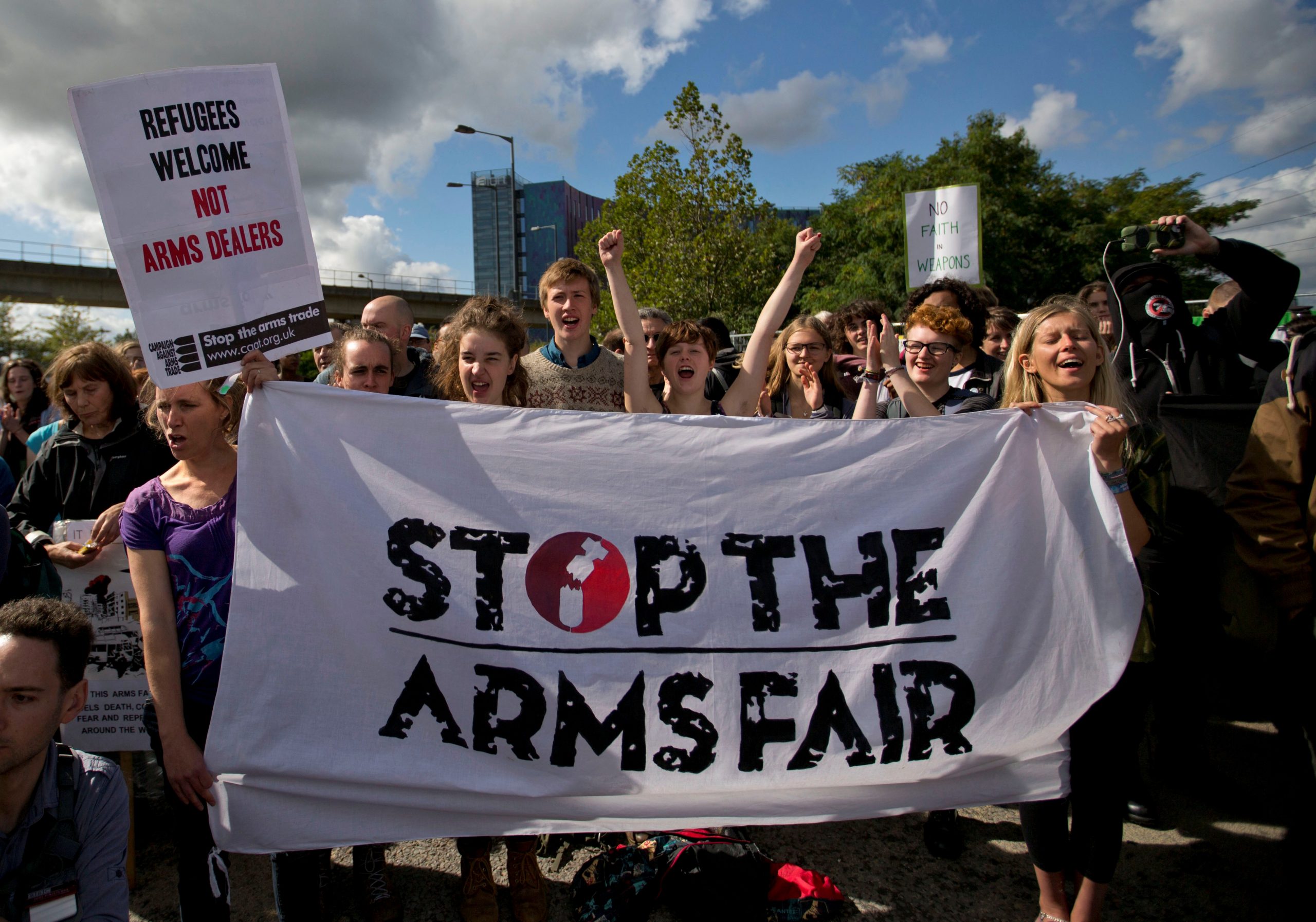A group of people stand behind a 'Stop the Arms Fair' banner chanting