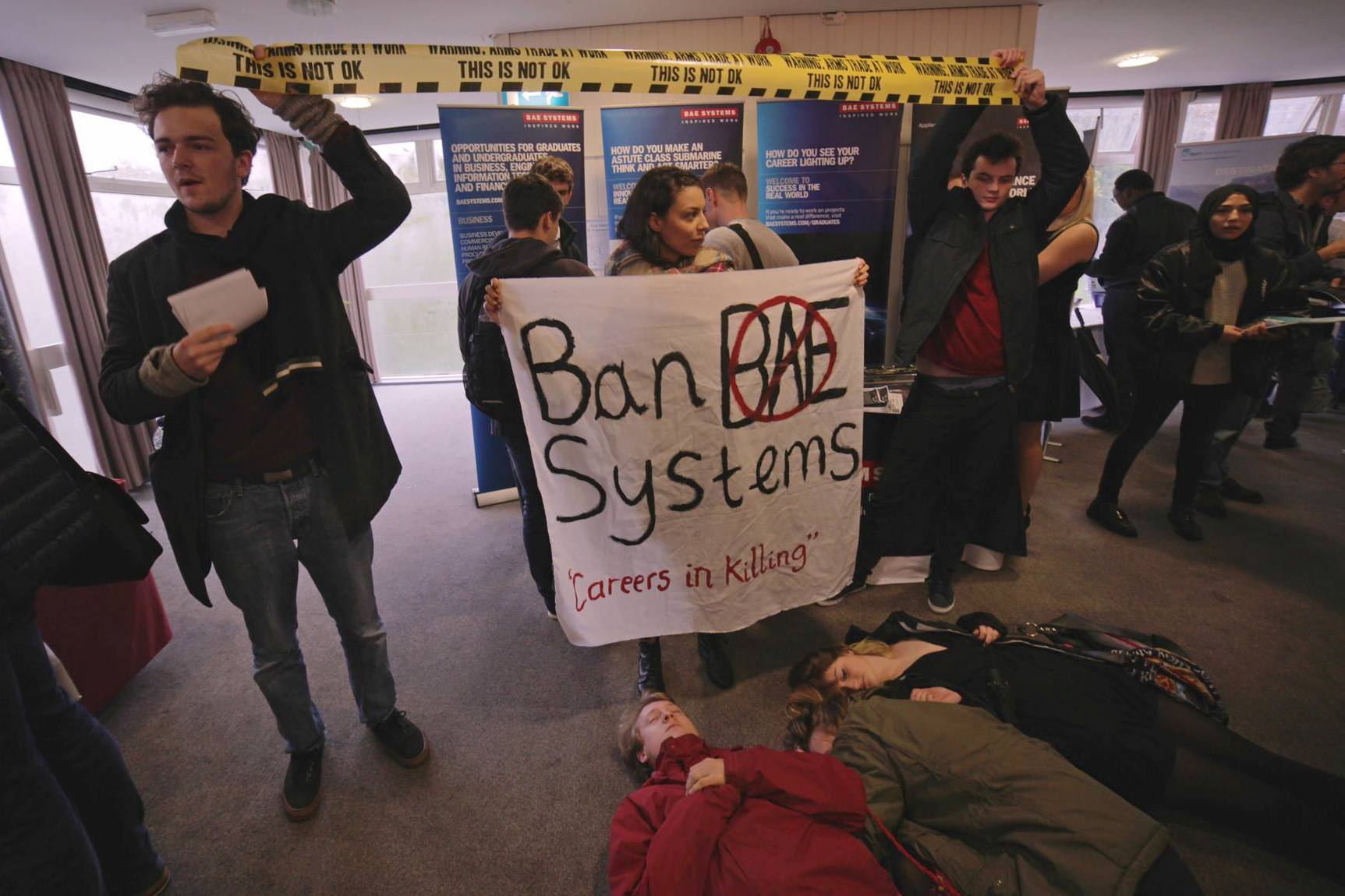A crowd of students stand in front of a BAE systems stall holding a "ban BAE Systems"banner and warning tape that reads "cautios, arms trade at work". Several students lie on the floor pretending to be dead.