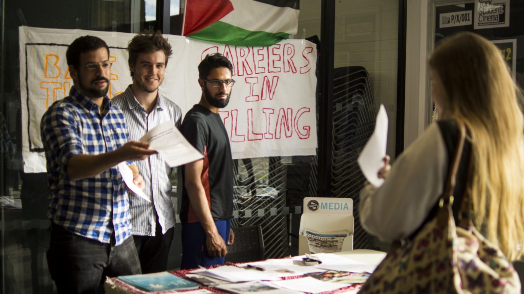 Three people hand out leaflets at an information stall. There is a banner that reads "careers in killing" in the background.