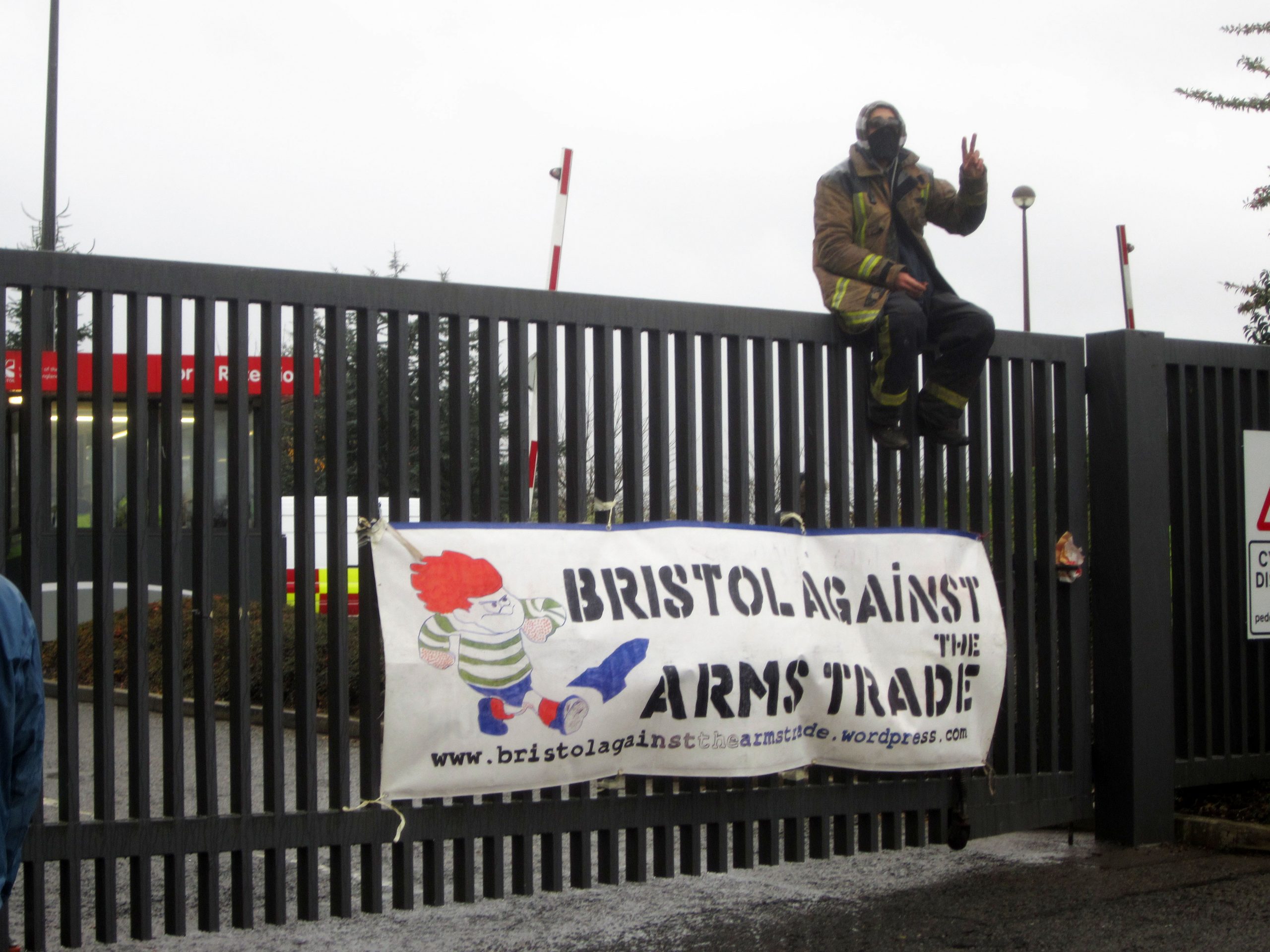 A protester sits on top of a closed gate to UWE with a &quot;Bristol Against the Arms Trade&quot; poster