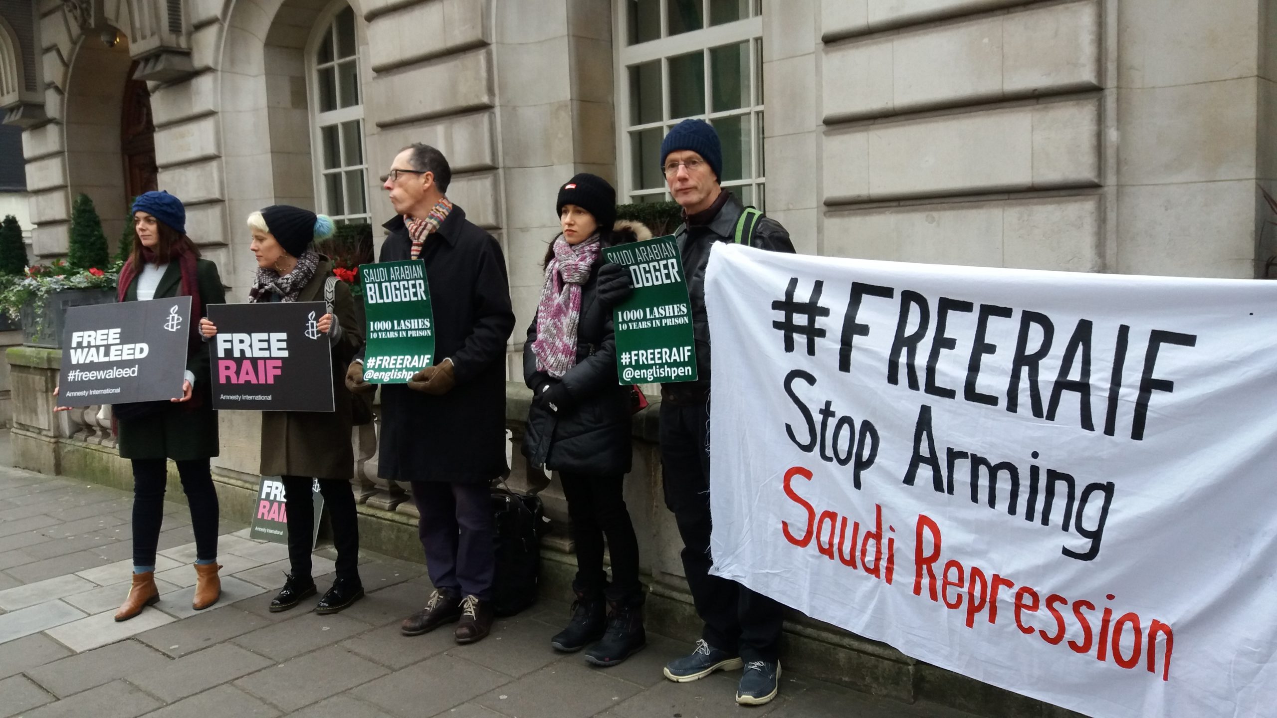 Protesters stand outside the Saudi embassy. They hold placards that read, “Free Raif”, “Free Waleed”, “Saudi Arabian blogger imprisoned - 1000 lashes - 10 years in prison - #FreeRaif @EnglishPEN” and a large banner that says “#FreeRaif, Stop Arming Saudi Repression”