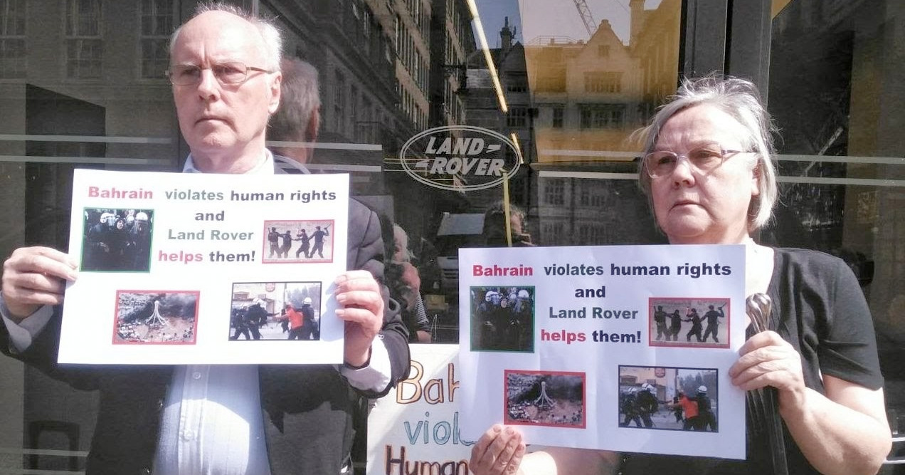 Two older white people with grey hair stand in front of a Land Rover logo holding posters that read 'Bahrain violates human rights and Land Rover helps them!'