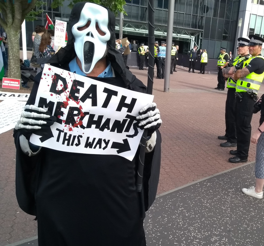 A protester dressed as 'death', holding a sign with arrows that reads 'Death Merchants this way'