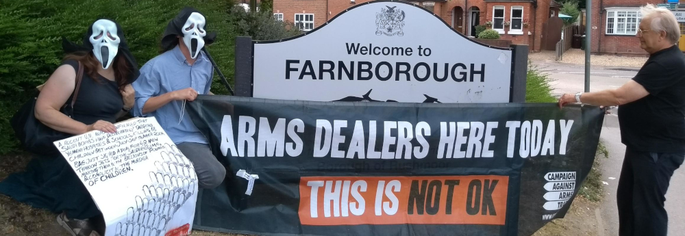 Protesters holding a banner in from of a 'Welcome to Farnborough' sign. The banner reads 'Arms Dealers Here Today'.
