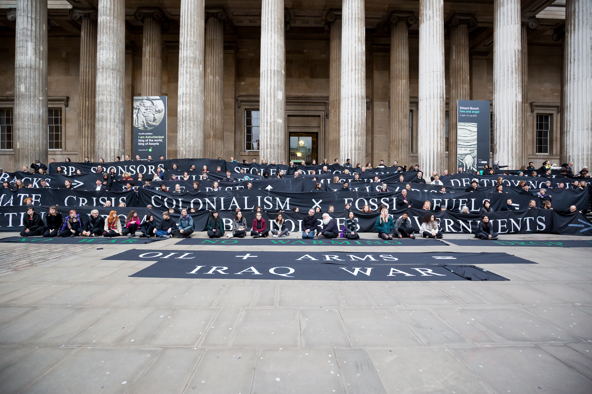 Protesters stand outside the British Museum in lines holding onto large banners/ a 'living tapestry' with messages about war, colonialism, the Iraq war and fossil fuels painted on