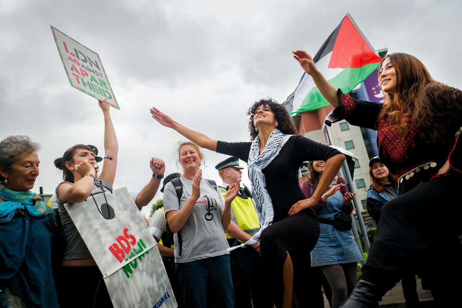 Protesters hold a sign and cheer on two dabke dancers with a policeman and Palestinian flag in the background