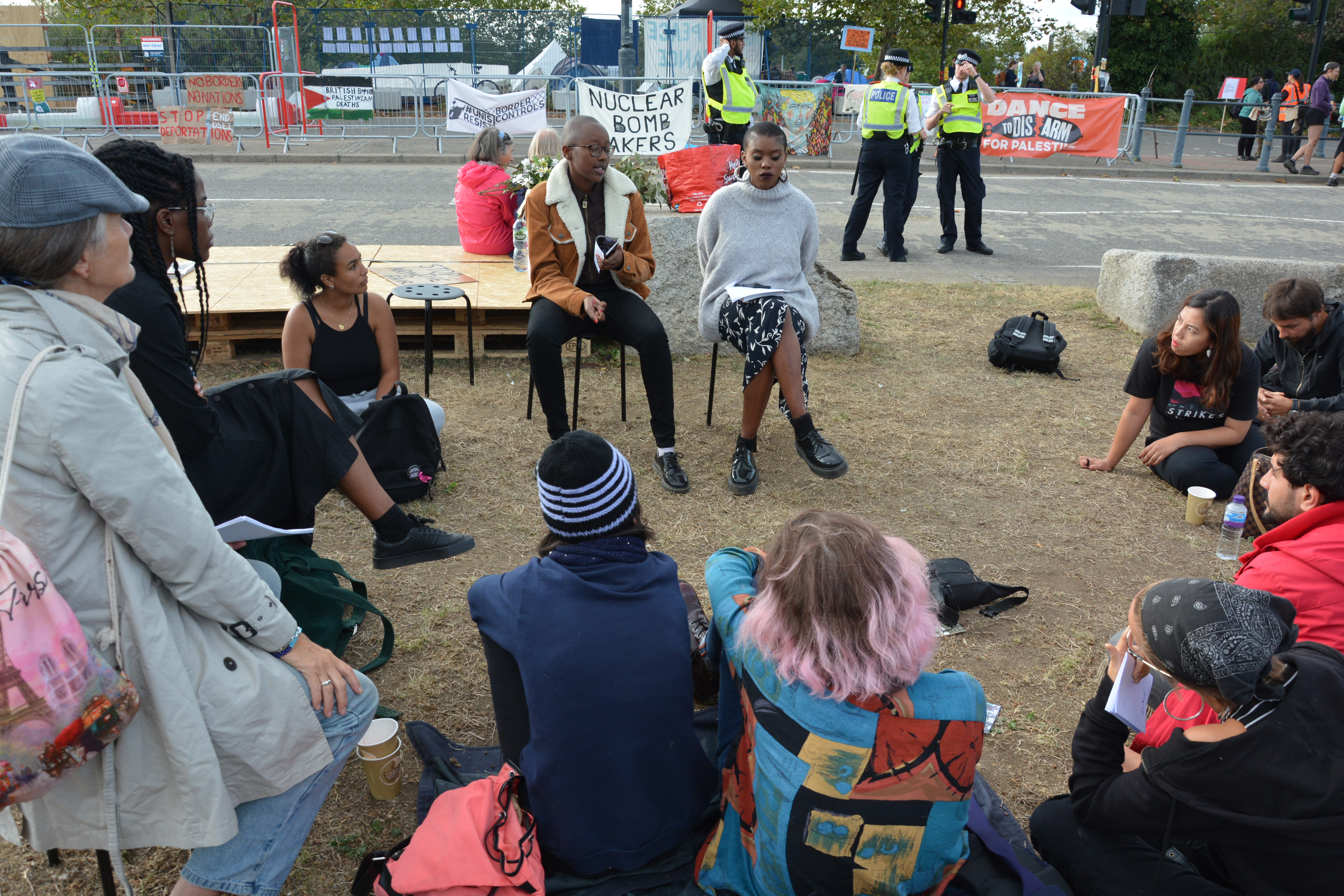 A group of people listening to someone speak, seated in a circle next to a road. Police are nearby