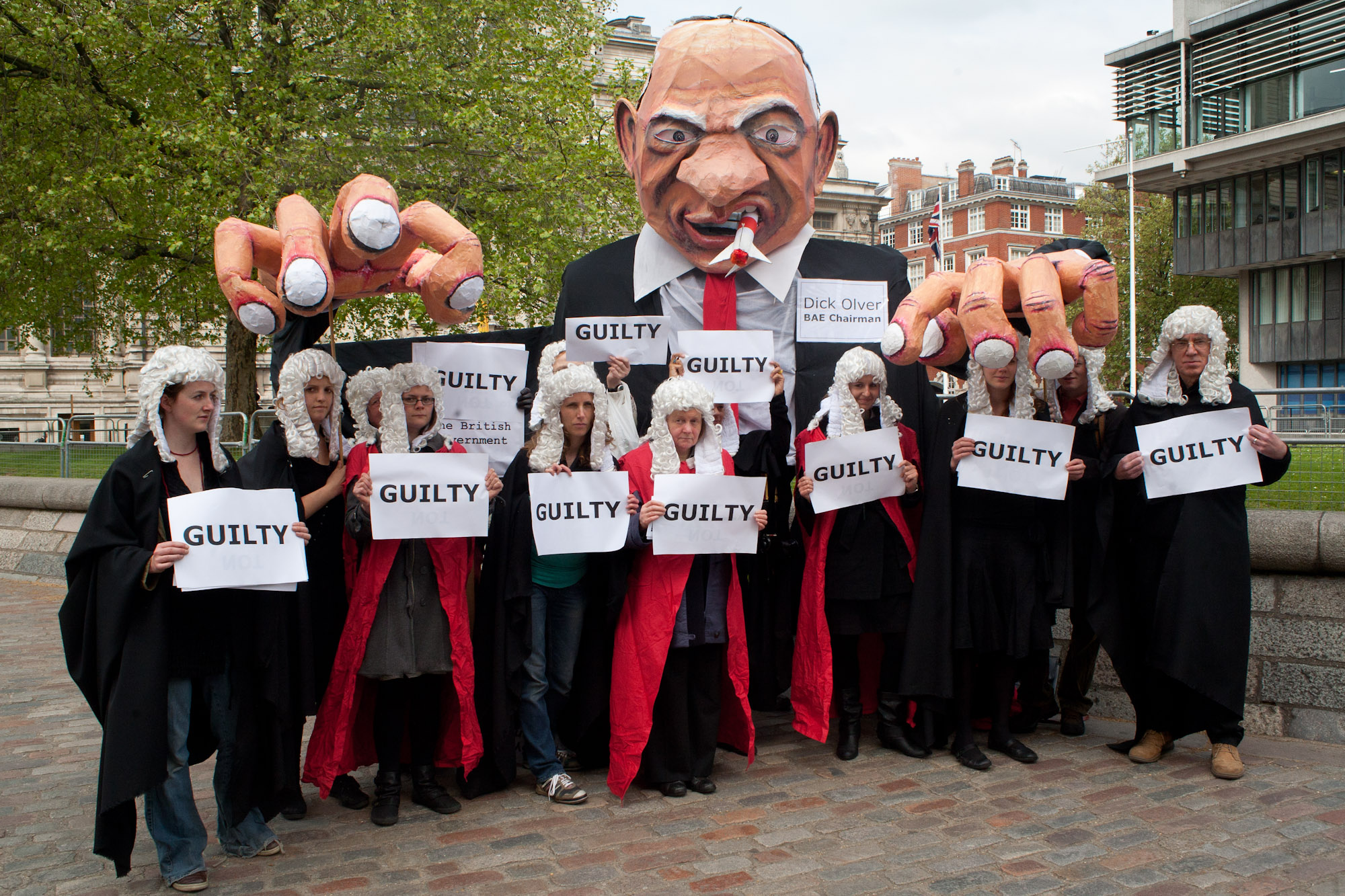 Giant paper mache puppet of man and 2 dozen people in foreground dressed as barristers with white paper reading &quot;Guilty&quot;