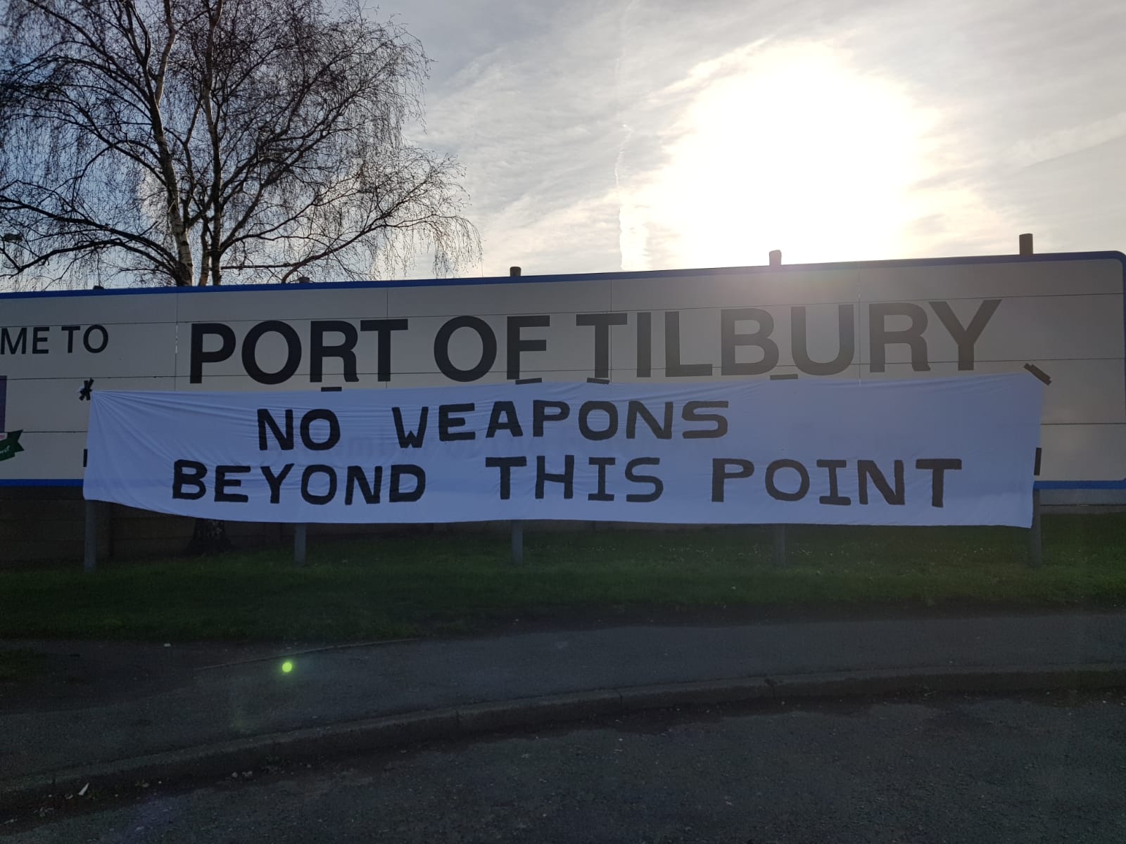 Sun in background with white banner reads &quot;No weapons beyond this point&quot; in foreground over sign reading &quot;port of tilbury&quot;