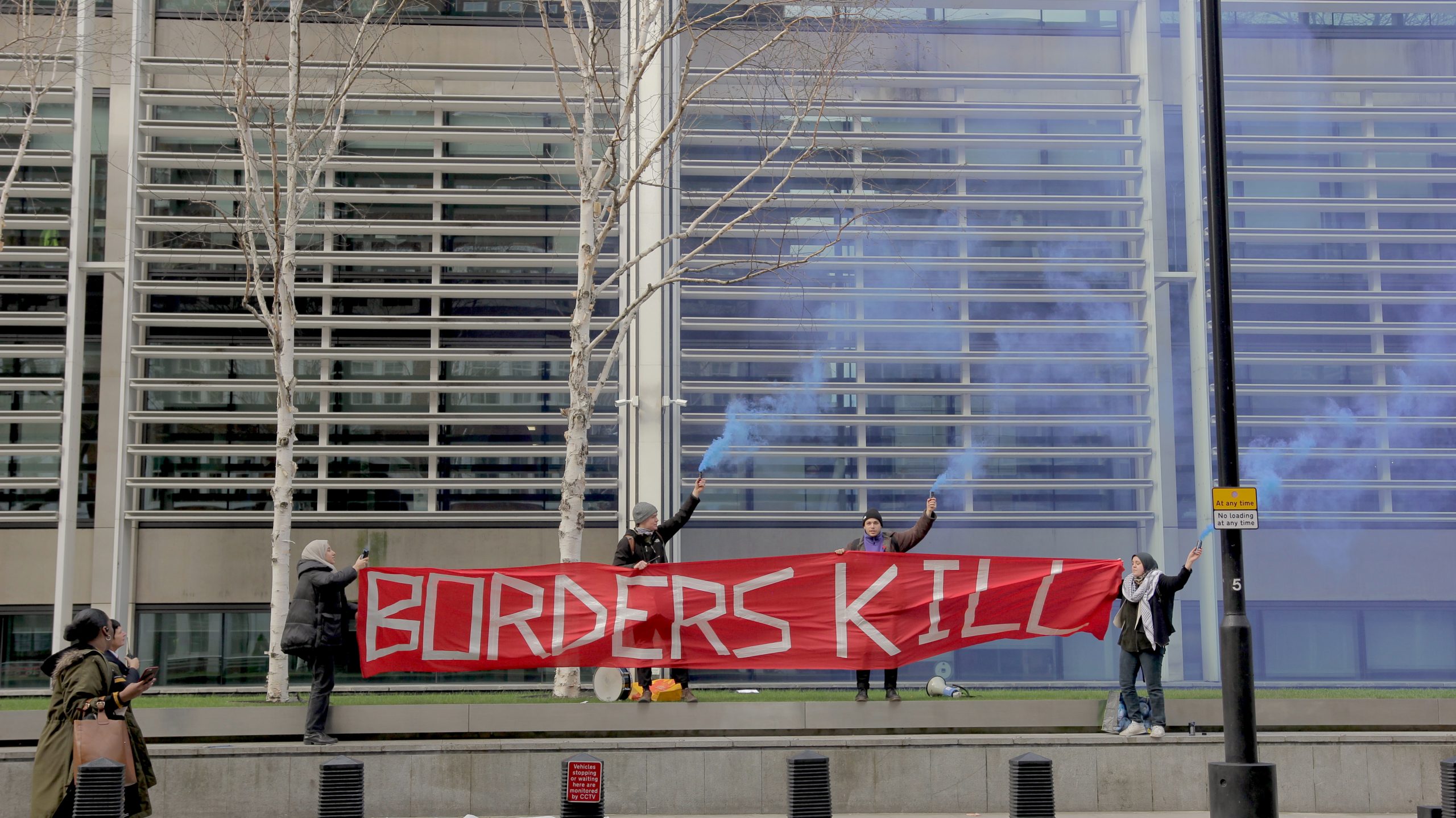 Protestors hold a big red banner that states &#039;Borders Kill&#039; and blue flares outside the Home Office