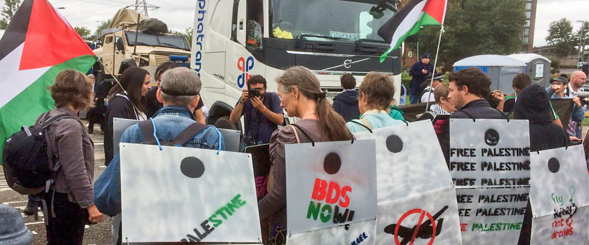 Crowd of activists standing in front of a blocked truck at an arms fair carrying signs saying &quot;BDS Now!&quot; and carrying Palestinian flags.