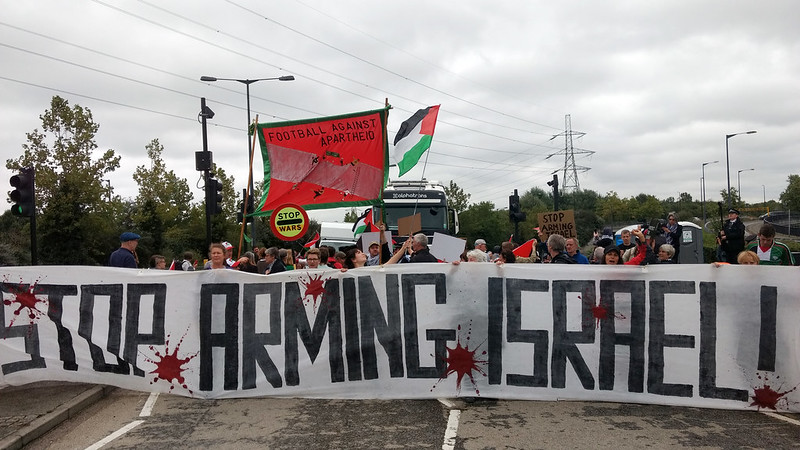 People holding a large banner across a road with a truck behind it. The banner reads 'Stop Arming Israel'