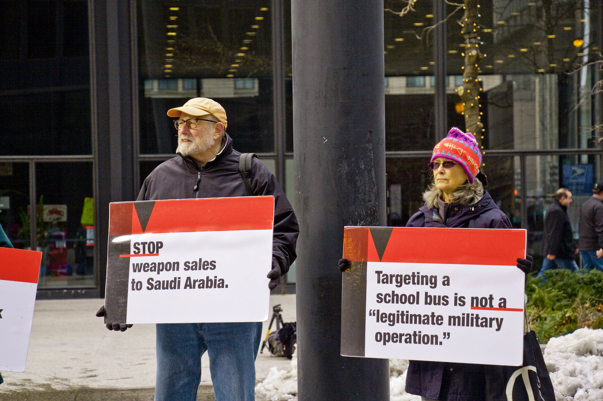 Signs Stop weapons sales to Saudi Arabia and Targeting a school bus is not a legitimate military operation; held by two activists, one wearing a beard and flat cap. The other wearing a pink and purple hat.