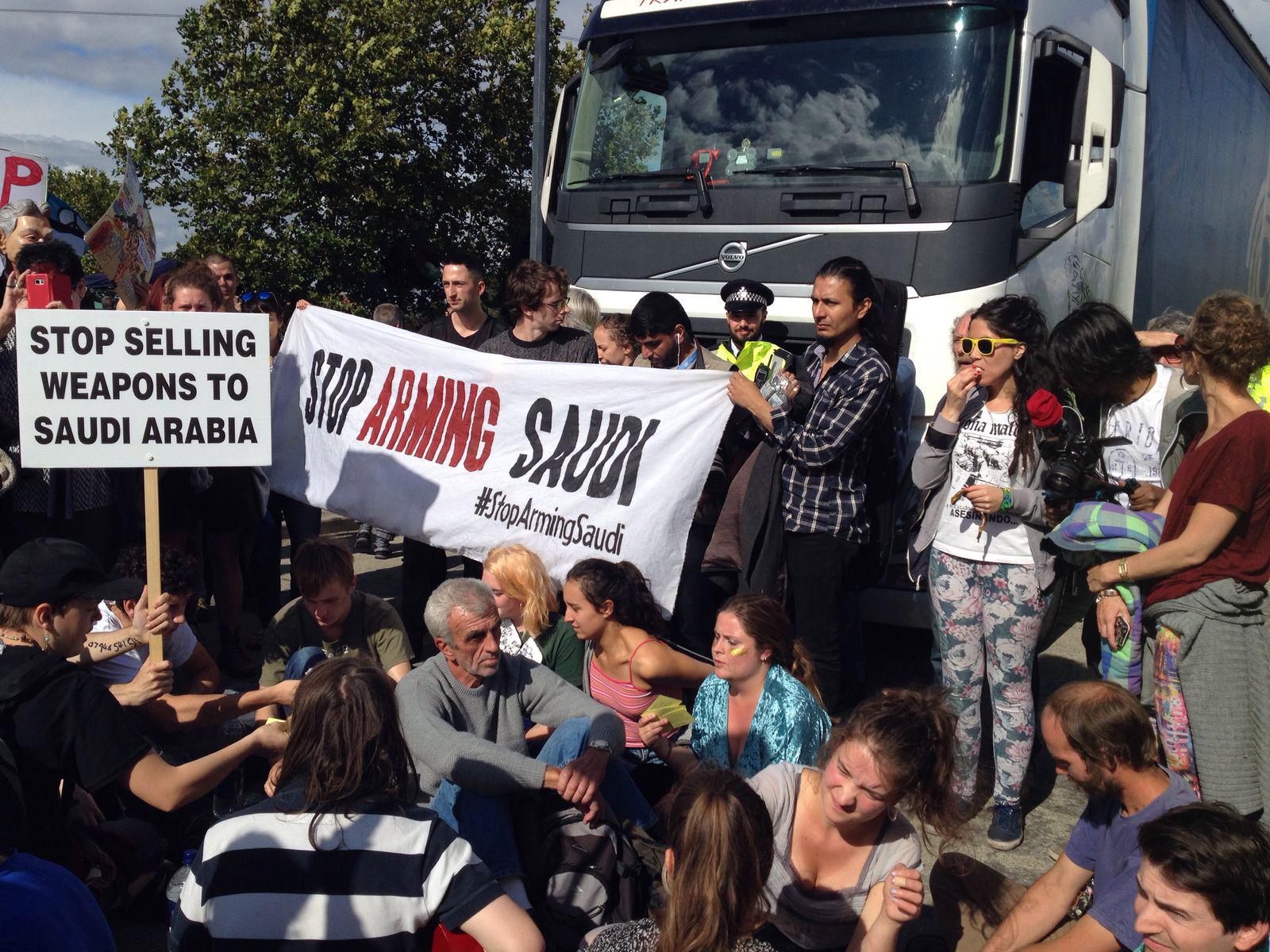 Stop Arming Saudi banner held by ten activists in front of a truck. Twenty activists sat in front of the banner. An activist with yellow sunglasses long dark hair and patterned leggings is eating it appears a piece of fruit watching on,.