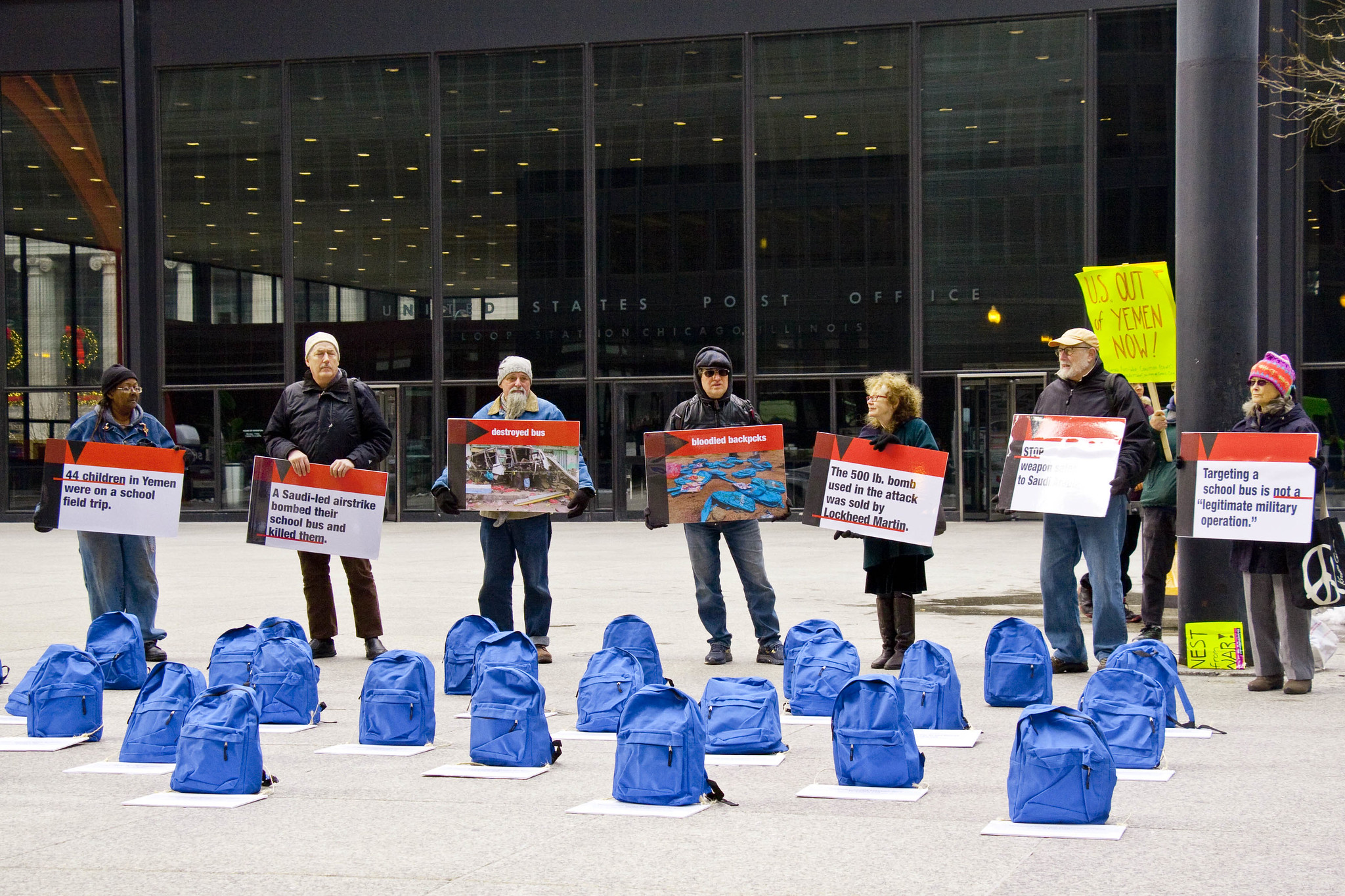 Seven campaigners holding signs standing behind 20 blue school bags. Signs are at a distance but appear to relate to Yemen and a school bus bombing