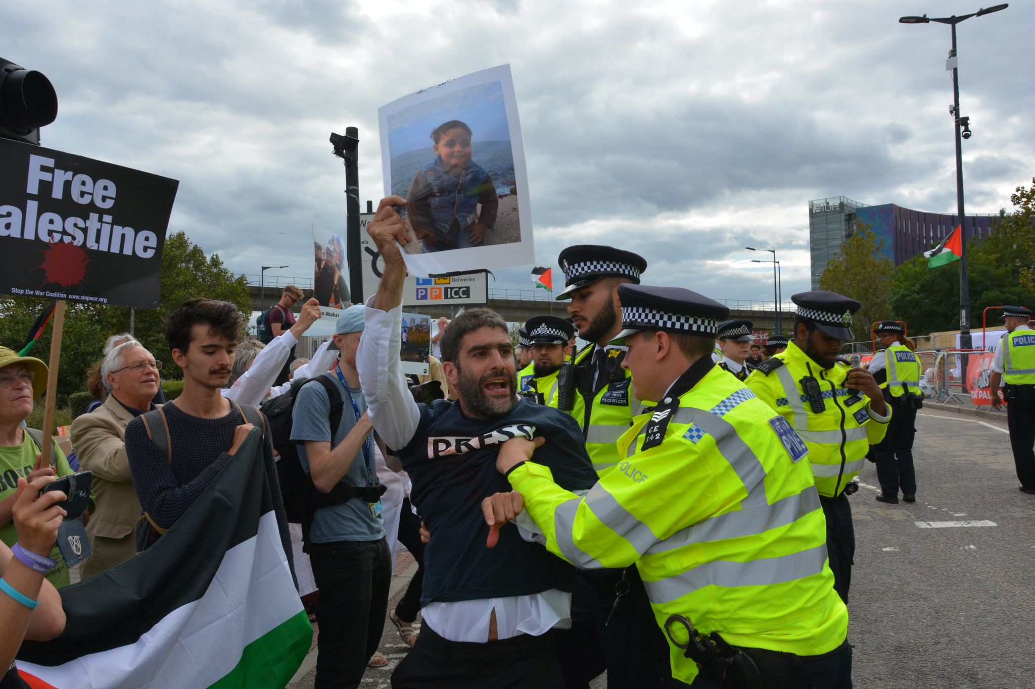 Bearded campaigner holding a picture of a child in front of a crowd carrying Palestine flags. They are shouting. 2 police officers are holding the campaigner with the picture back from a road going into Excel in London.