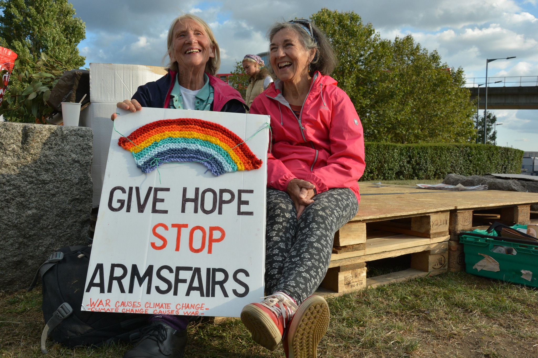 Give Hope Stop Arms Fairs sign with woven rainbow held by two campaigners, one wearing a pink jacket, the other wearing a black and pink jacket.