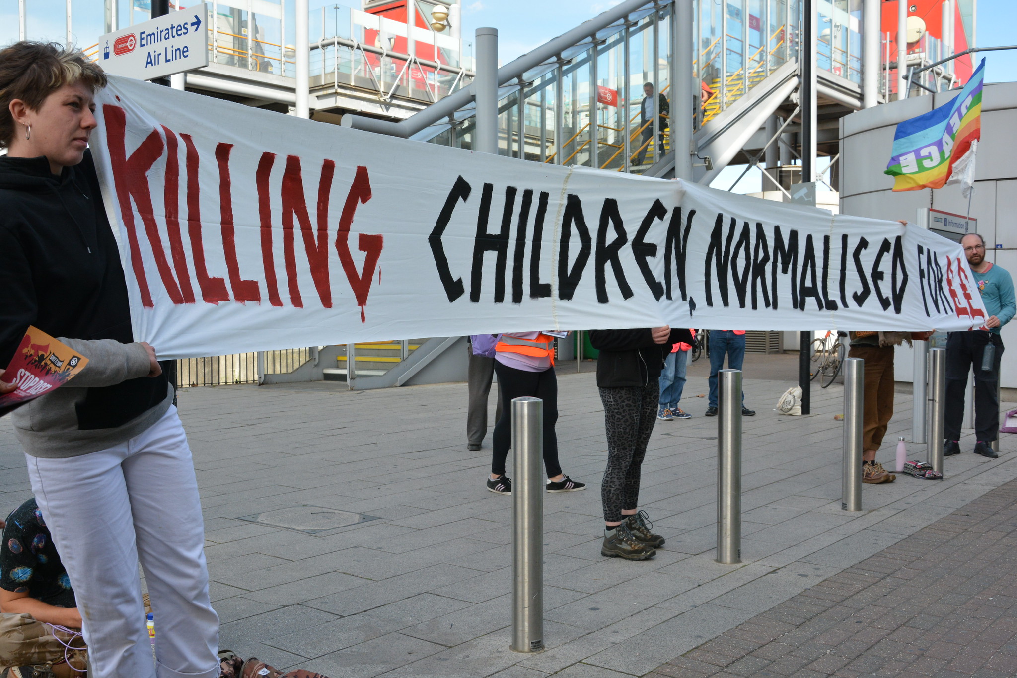Campaigners at Royal Victoria DLR holding a sign saying Killing Children Normalised for££