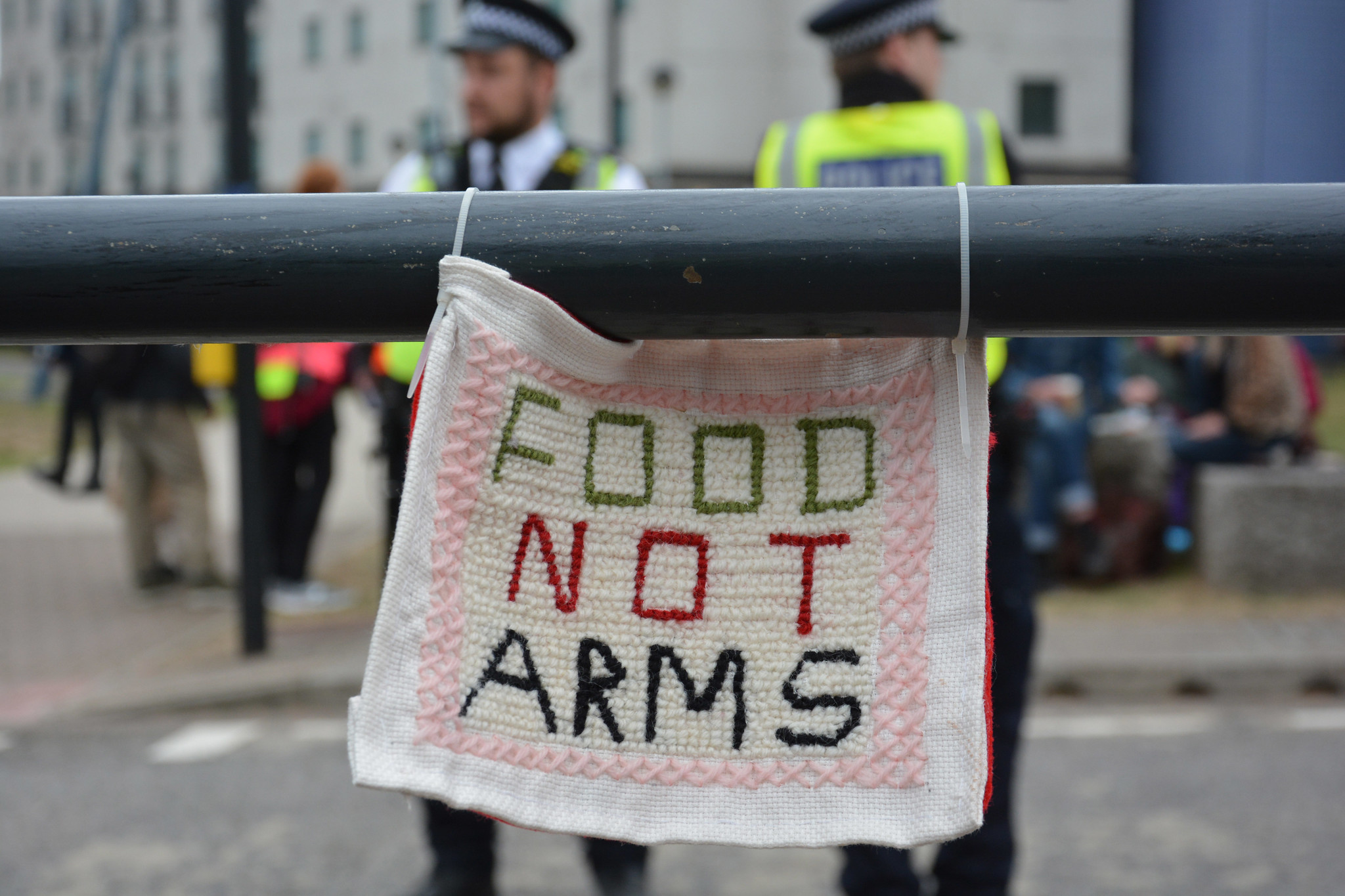 Food Not Arms embroidered banner hanging from a pipe; cops out of focus behind