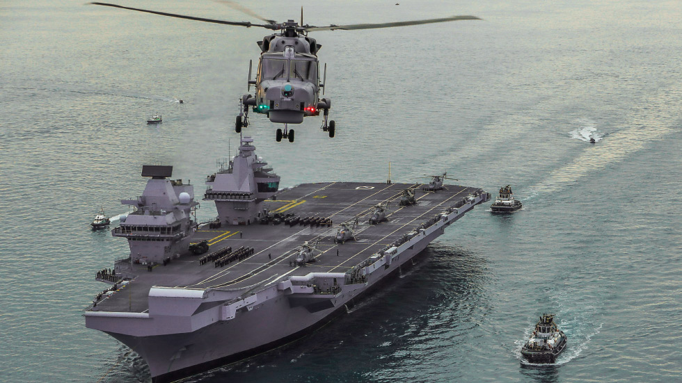 HMS Queen Elizabeth class carrier with complement of helicopters and sailors standing in rows on deck. Tugs escorting. Helicopter flying in foreground with green and red lights