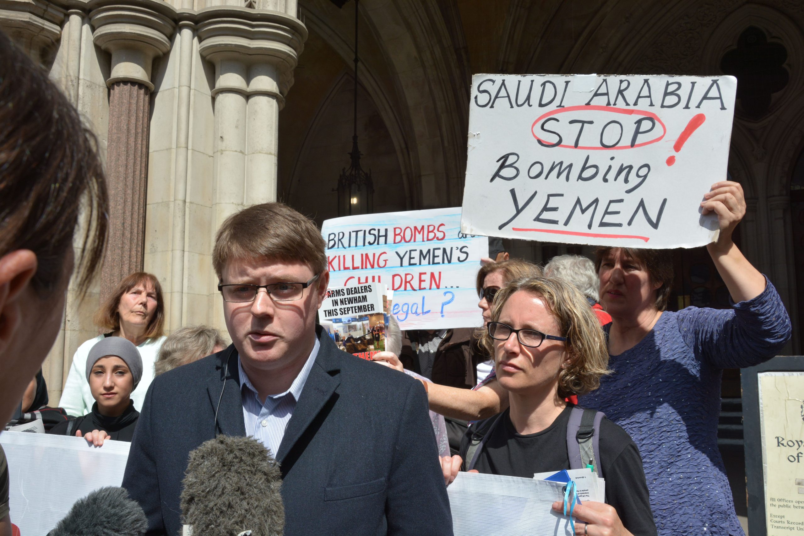 Andrew Smith from CAAT pictured outside the Royal Courts of Justice surrounded by activists holding signs saying Saudi Arabia Stop Bombing Yemen and British Bombs Killing Yemens Children. Andrew is talking to a reporter carrying a broadcast microphone