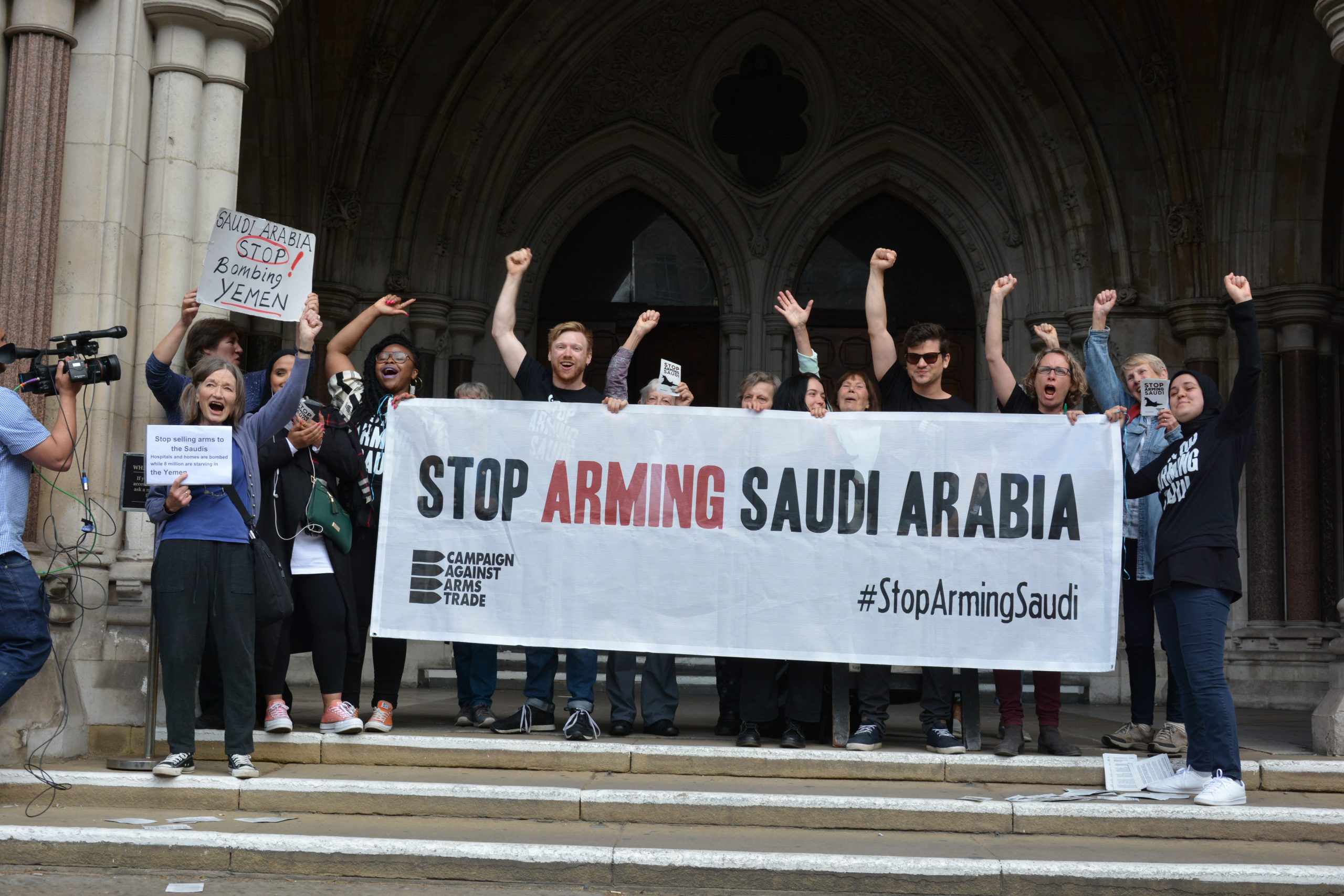 Campaigners with a Stop Arming Saudi banner outside the Royal Courts of Justice, another holds a sign saying Saudi Arabia Stop Bombing Yemen. They all appear happy and are raising their fists in the air.
