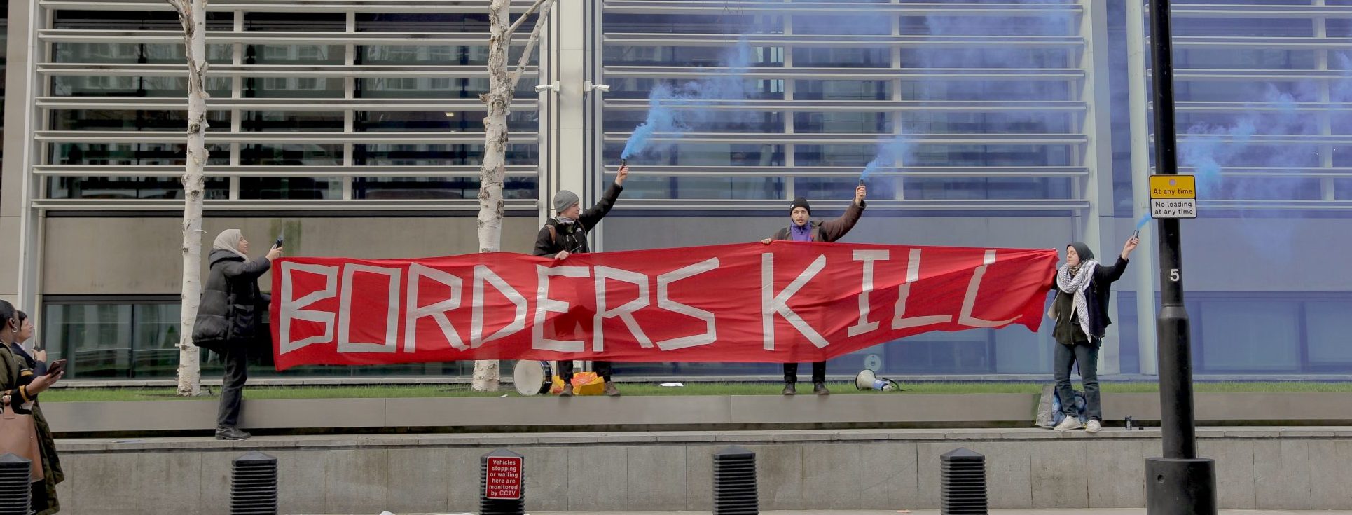Borders Kill banner carried by four campaigners with hats and headscarfs outside Home Office. Three hold smoke pyrotechnics