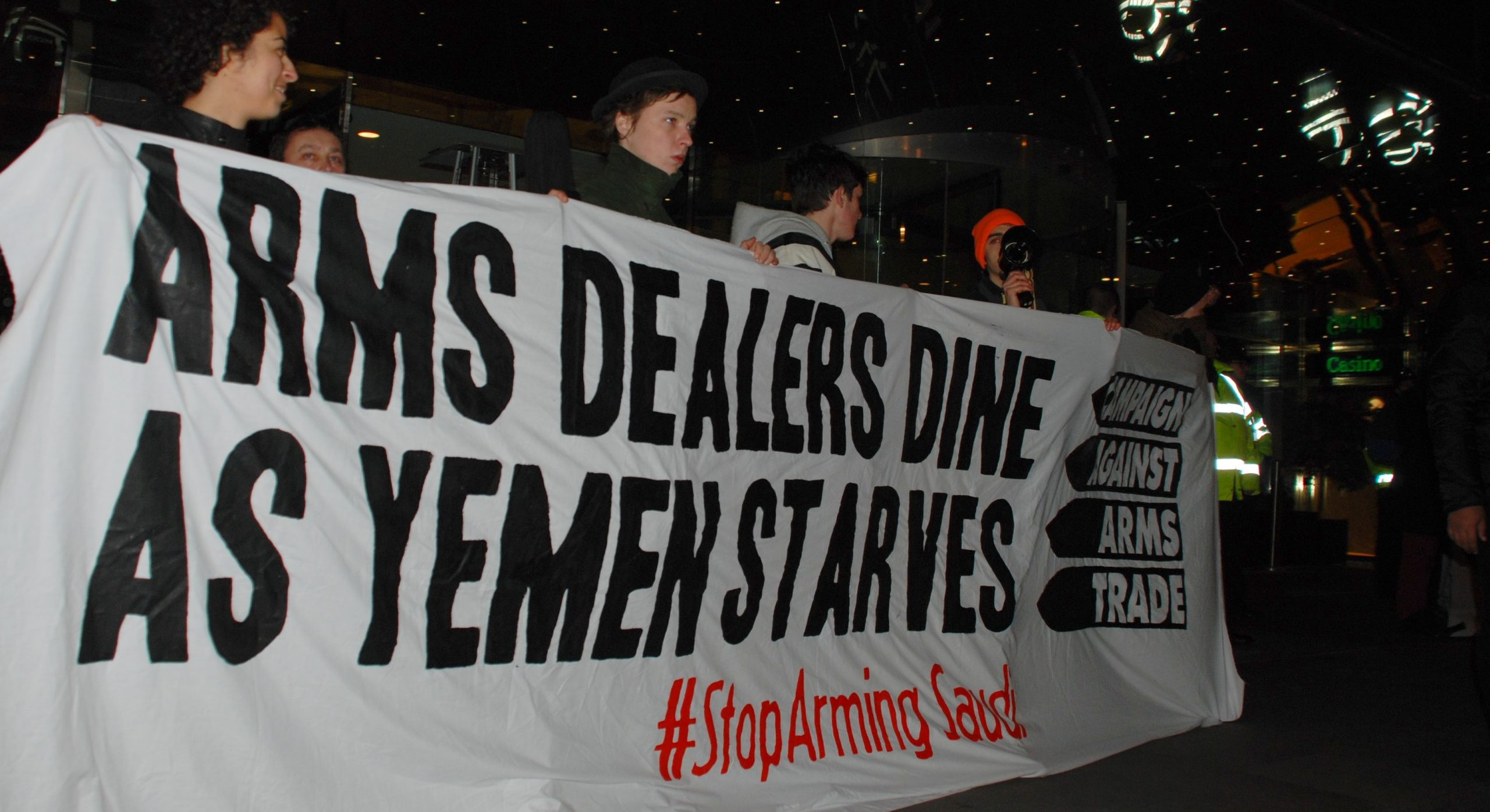 Arms Dealers Dine as Yemen Starves #StopArming Saudi CAAT banner held by four activists outside a hotel under a mirrored canopy. The mirror reflects policeman in fluorescent jackets.