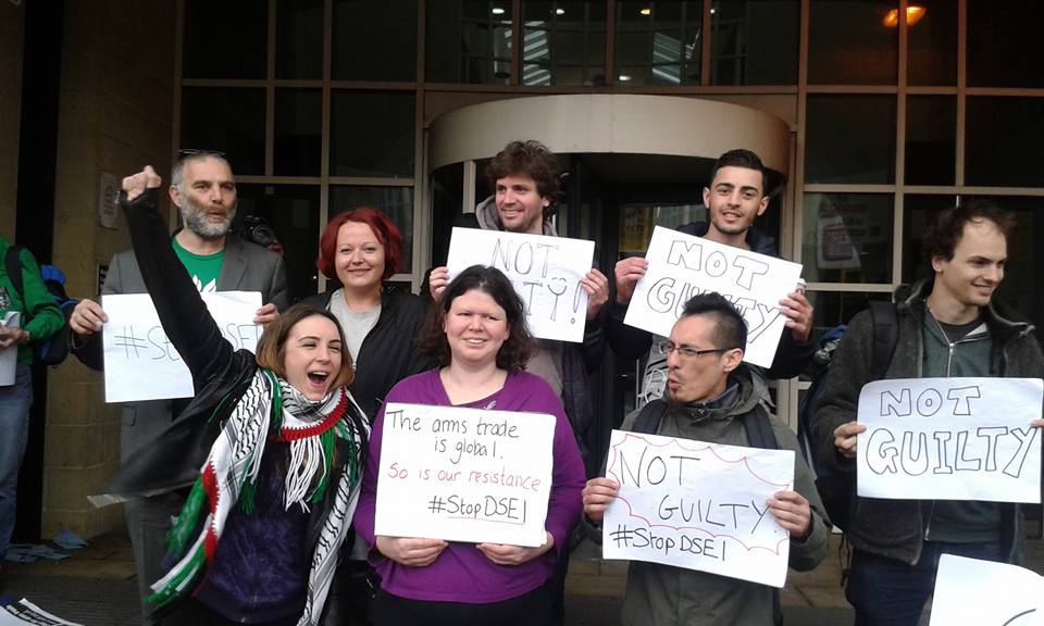Image of seven people, three in front four in back, in front of a court building, holding placards mostly saying &quot;Not Guilty&quot;, one also saying &quot;#StopDSEI&quot;, another saying &quot;The arms trade is global, so is our resistance, #StopDSEI&quot;. Woman at front on left wearing a Palestinian scarf, raising clenched fist and cheering. Court building behind them.