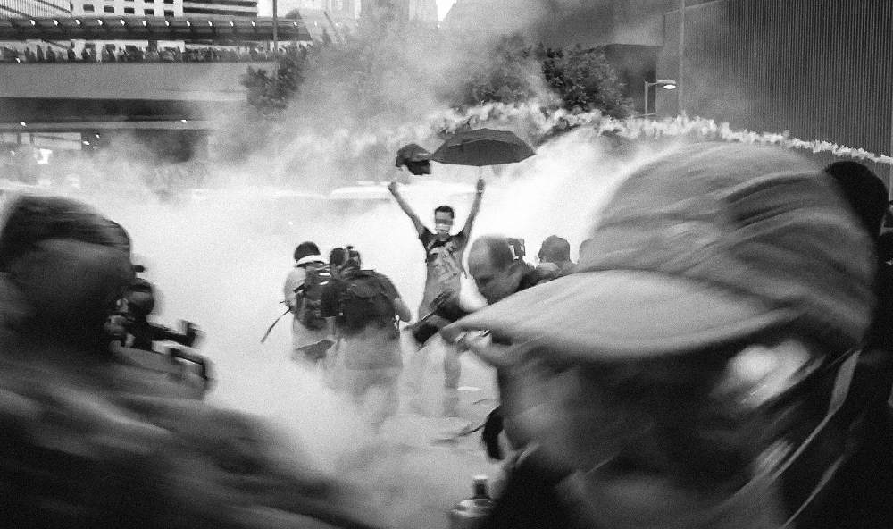 Protesters in a cloud of tear gas. One in the middle with raised arms, holding an umbrella