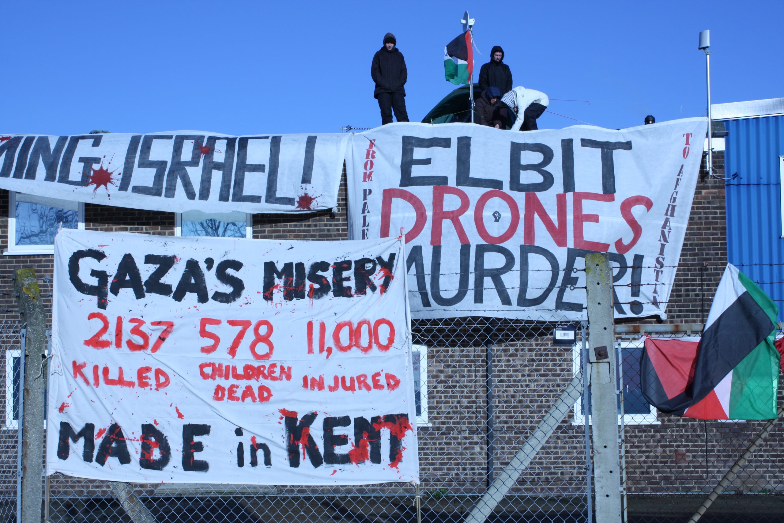 Activists stand on a factory roof behind barbed wire. Banners read 'Stop arming Israel!', 'Elbit drones murder!', and 'Gaza's misery; 2137 killed, 578 children dead, 11,000 injured; Made in Kent'
