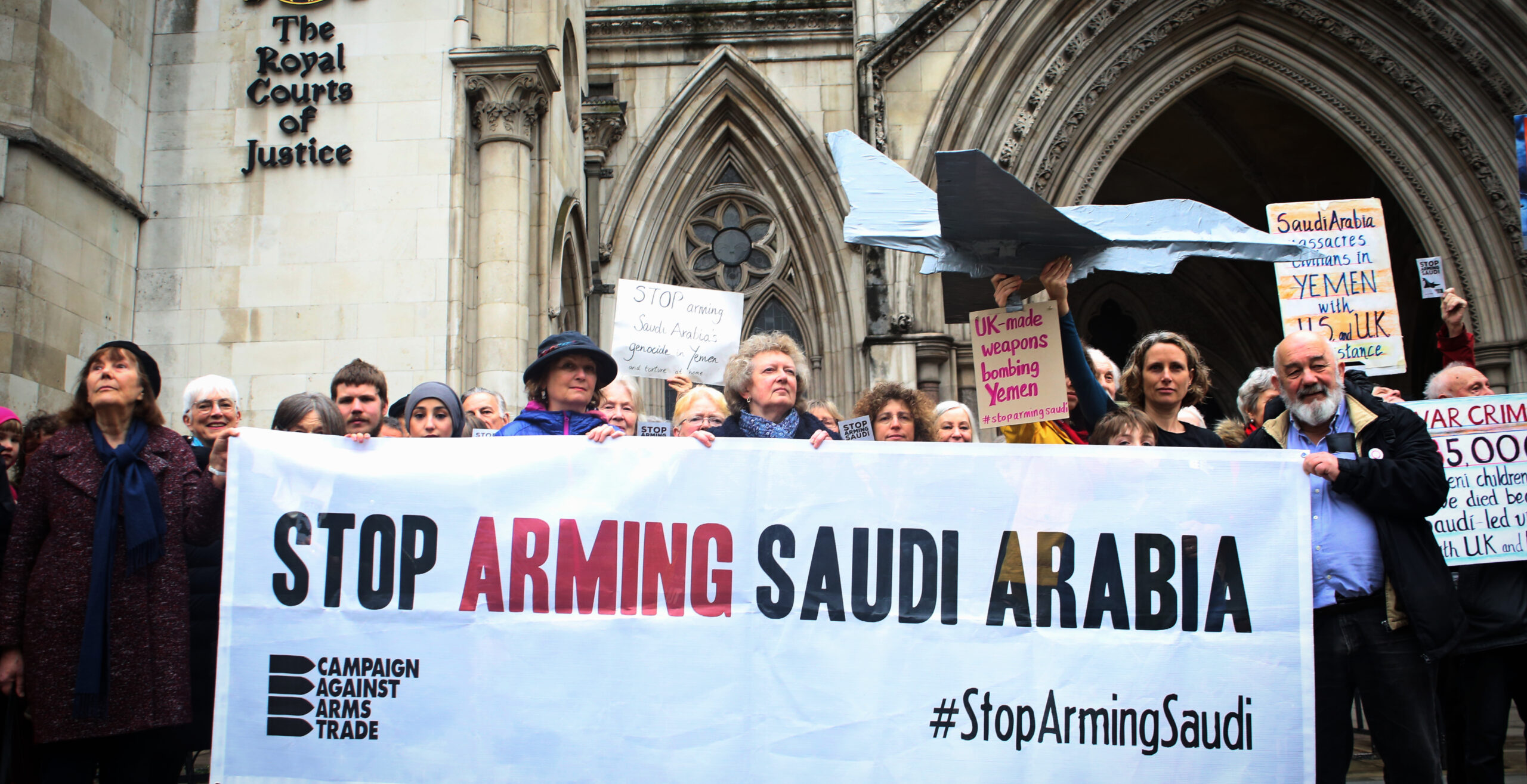 crowd of people standing in front of the Royal Courts of Justice with a 'Stop Arming Saudi Arabia #StopArmingSaudi, Campaign Against Arms Trade' banner