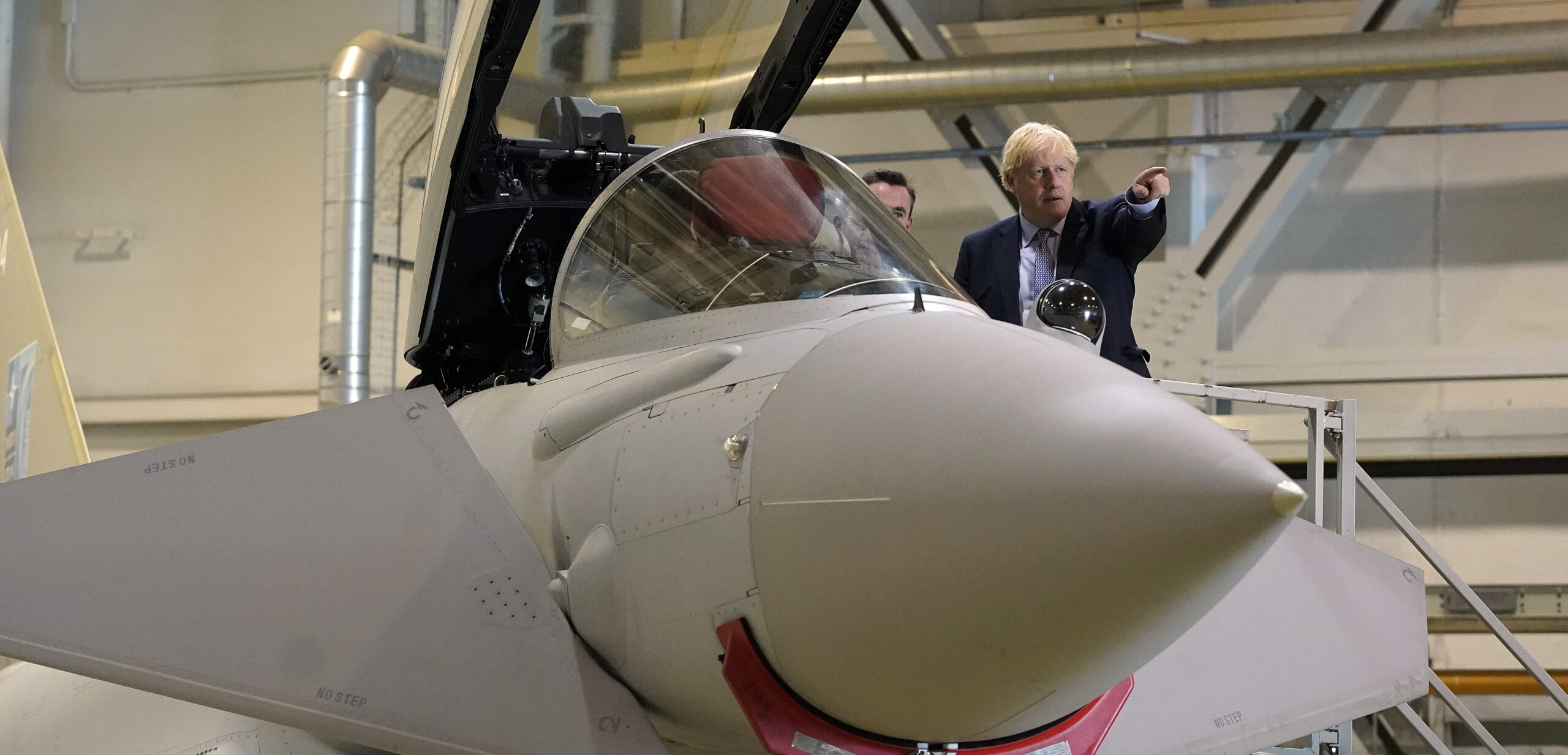 Boris Johnson standing next to a pilot sitting in Typhoon cockpit in a hangar. Johnson points to his left.