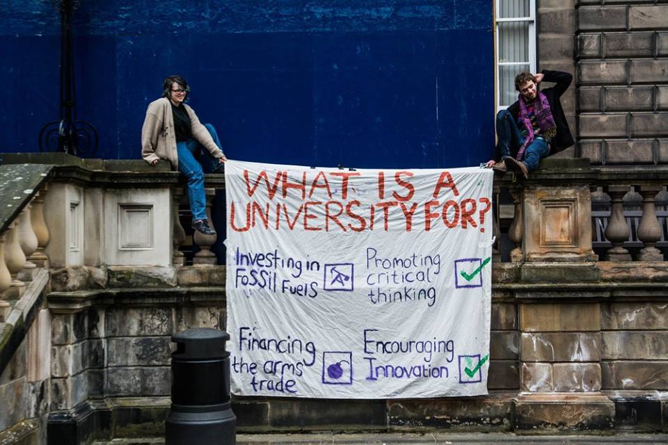 two students on building holding banner &quot;what is a university for?&quot; &quot;financing the arms trade&quot;
