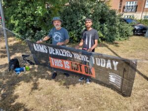 Two men hold banner saying, "Arms Dealers here today this is not OK"
