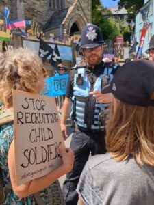 A protester at Armed Forces Day with a sign reading Stop Recruiting Child Soldiers alongside a Police Liaison Officer
