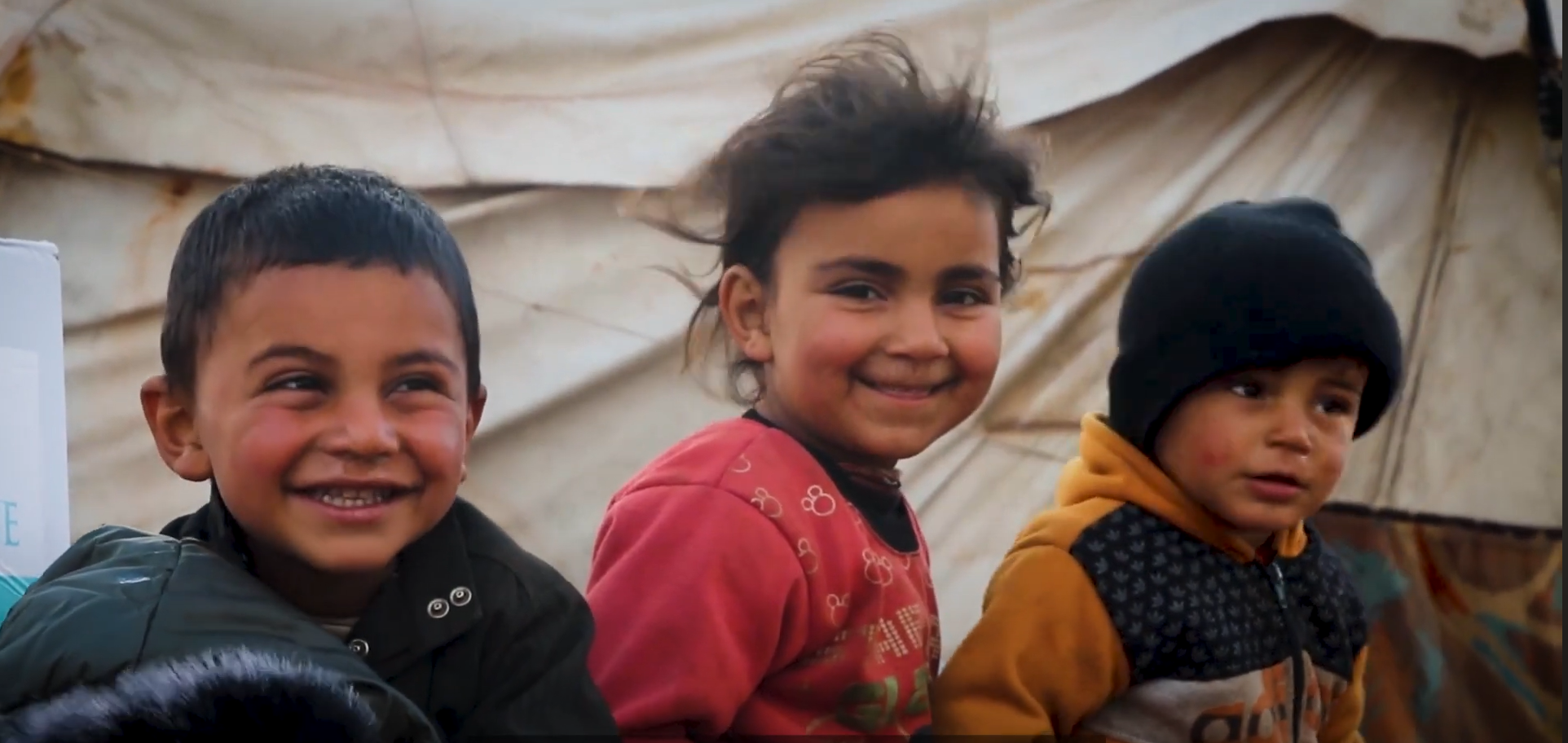 Three children next to a UN-style white tent. They are smiling and brightly clothed.