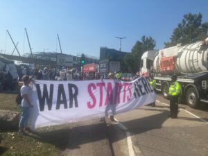 Protesters in road blocking lorries and holding banner saying "war starts here". Police in attendance