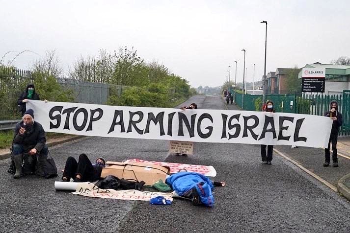 Protesters lying in the road locked together and other protesters standing above them with a stop Arming Israel banner