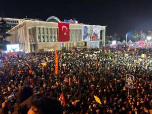 A large crowd with banners and posters outside a large plain concrete building draped with a large Turkish flag and large banners showing a male figure.