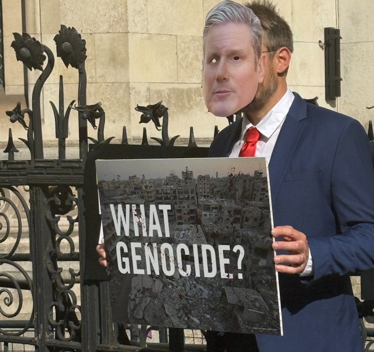 Photo of someone in a Keir Starmer mask with a sign saying what genocide outside the Royal Courts of Justice.