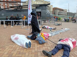 Death stands looking over a die in, as campaigners covered in bloodied shrouds lie on the ground
