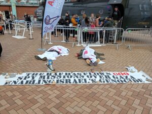 Campaigners dying in front of a Chinook helicopter giving tours to families and young people. They are covered in blood covered shrouds and have a banner that says, "The RAF has flown 600 spy missions over Gaza enabling Israel's Genocide"